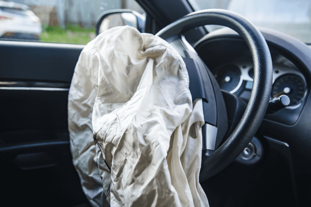 A deployed airbag, crumpled and white, hangs over the steering wheel inside a car. The interior is dimly lit, suggesting an outside environment with blurred greenery visible through the window.