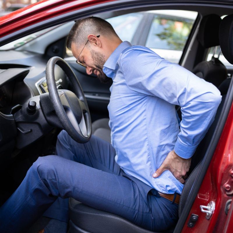 Man in car experiencing back pain while sitting, wearing a blue shirt and glasses, in a red vehicle.