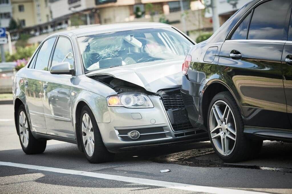 Two cars collided at an intersection, with visible damage to the front end and a shattered windshield on the silver car.