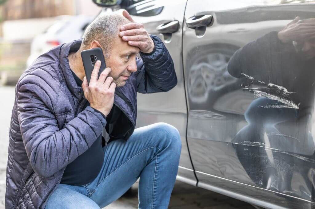 A distressed man uses a phone next to a car with a scratched door, indicating he’s likely reporting an accident or damage.