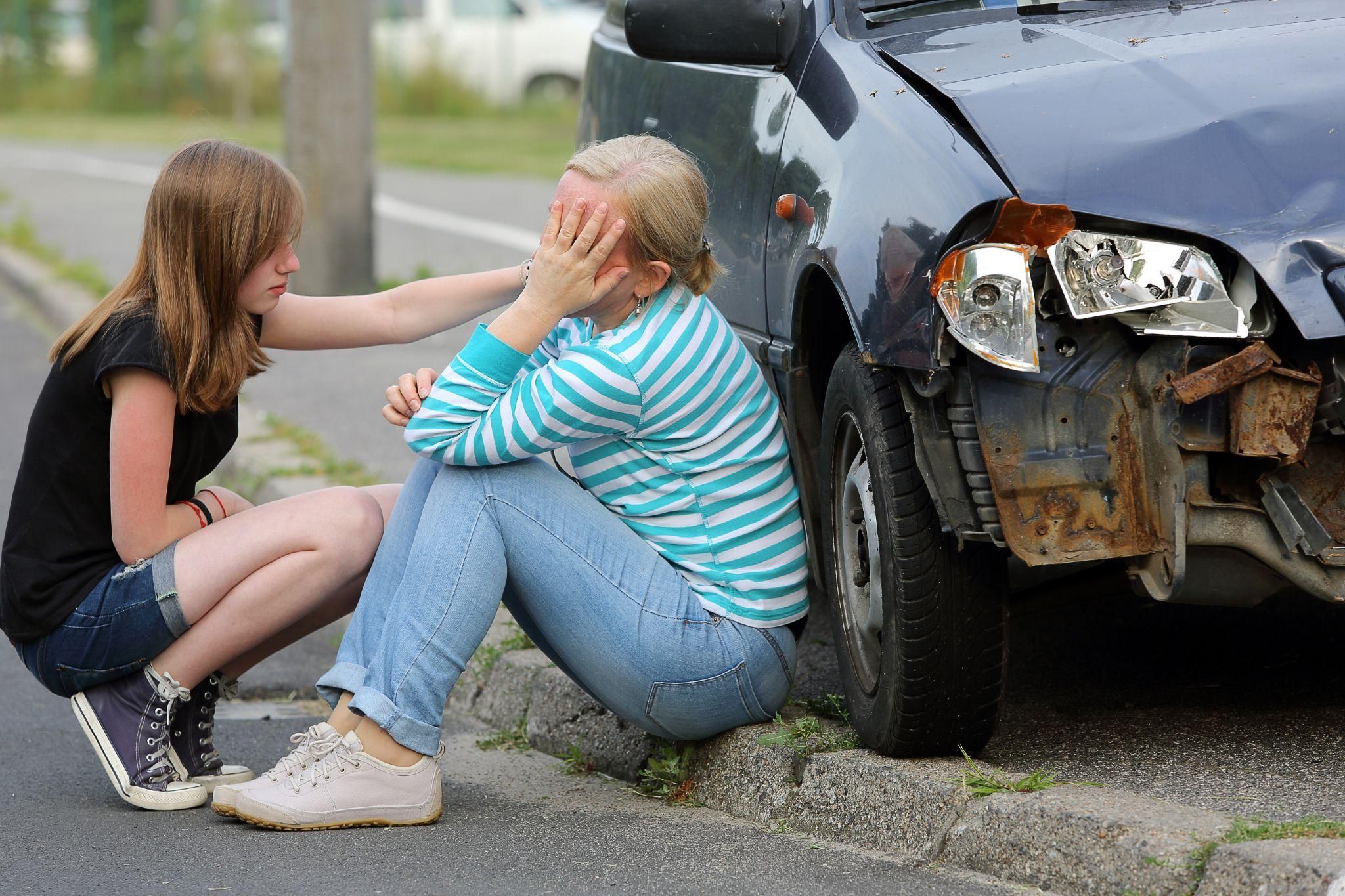 Teen comforting woman after minor car accident, damaged vehicle visible on roadside.