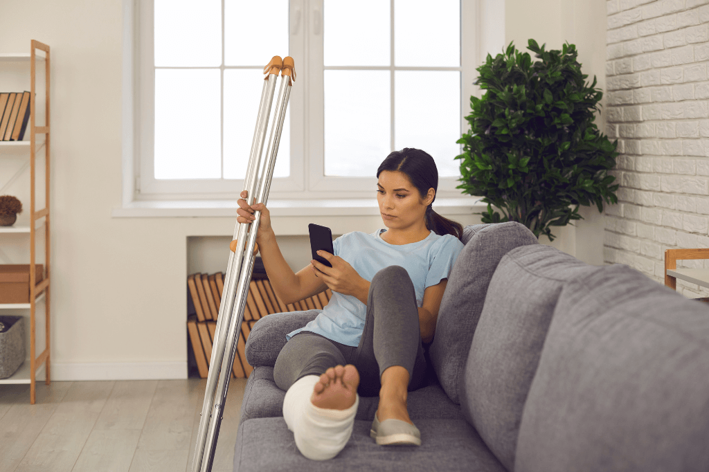 A woman sits on a gray couch, using a smartphone while resting her injured foot, wrapped in a bandage, on a nearby cushion. Crutches lean beside her in a bright, cozy room.