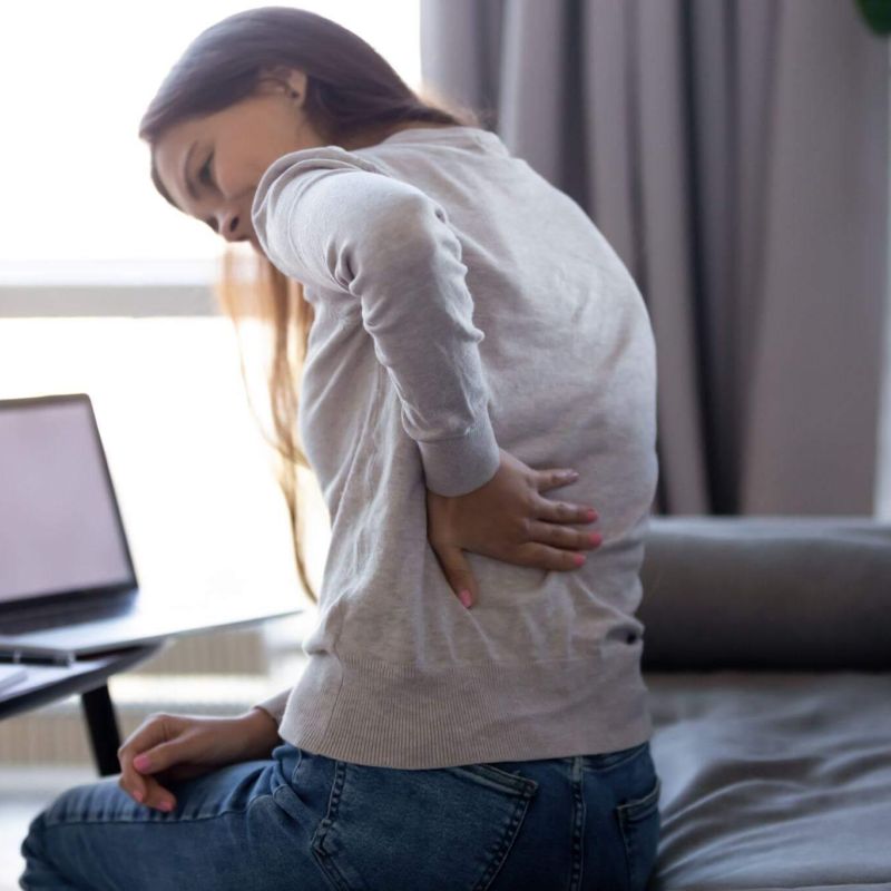 A person holding their lower back in discomfort, seated on a sofa next to a table with a laptop, in a bright and cozy living room.