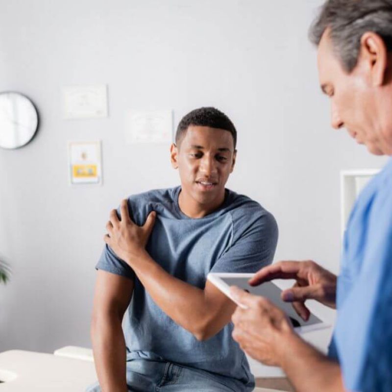A healthcare professional is using a tablet while examining a young patient's shoulder in a clinical setting with a clock and certificates on the wall.