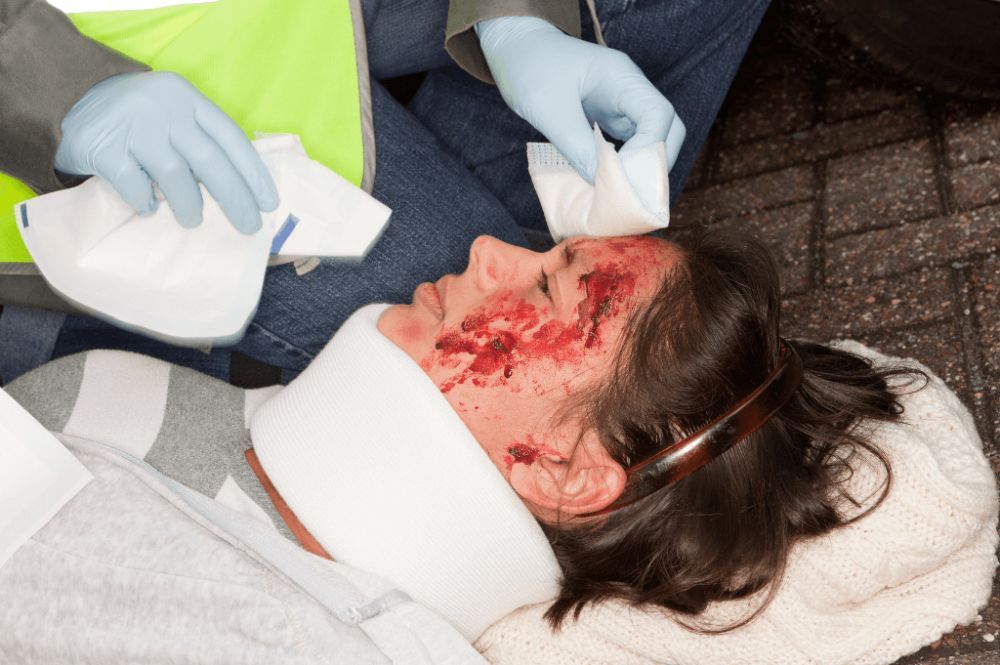 Emergency responder assists injured person with head wound and neck brace on pavement.