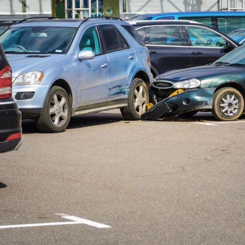A blue SUV is wedged against a green sedan, which has its front bumper partially dislodged. This accident occurs in a busy parking lot filled with various parked vehicles.