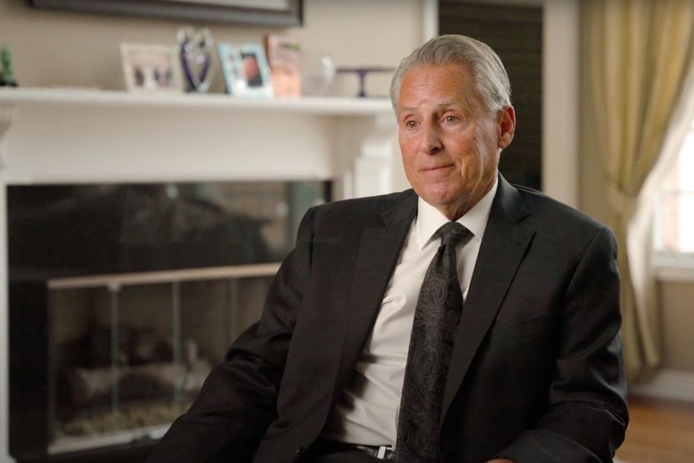 Man in a suit sitting in a well-decorated room, with a fireplace in the background.