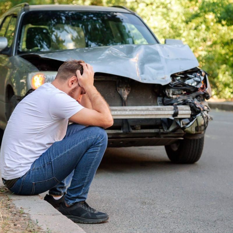 a man sitting on the side of a road next to a car, it seems they were recently involved in a car accident