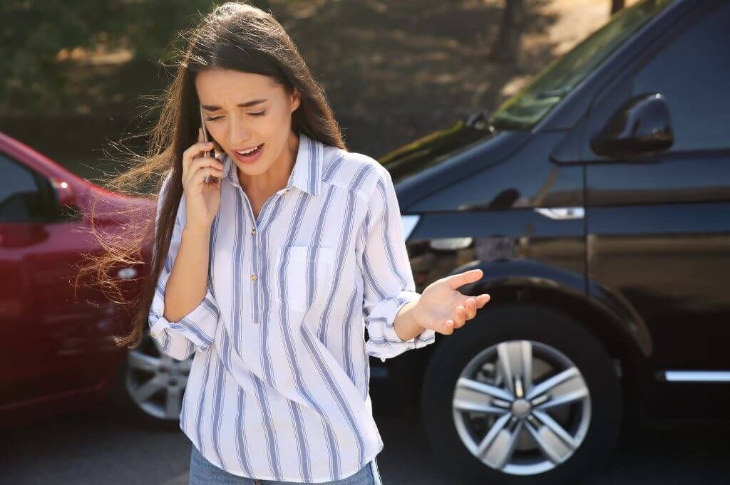 A woman standing outside her car, speaking on her phone with a distressed expression, in front of two vehicles that appear to have been involved in a minor collision.