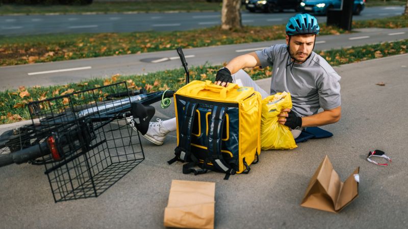 Cyclist in blue helmet sits on ground beside fallen bike, gathering items from a yellow delivery bag. Road in background.