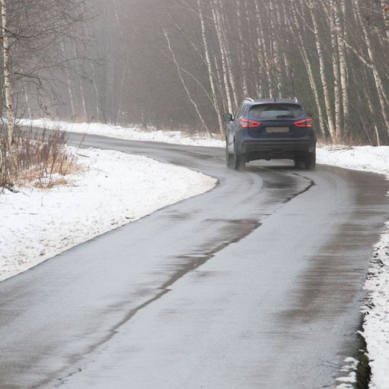 A car driving away on a winding wet road bordered by snow-covered ground and leafless trees.