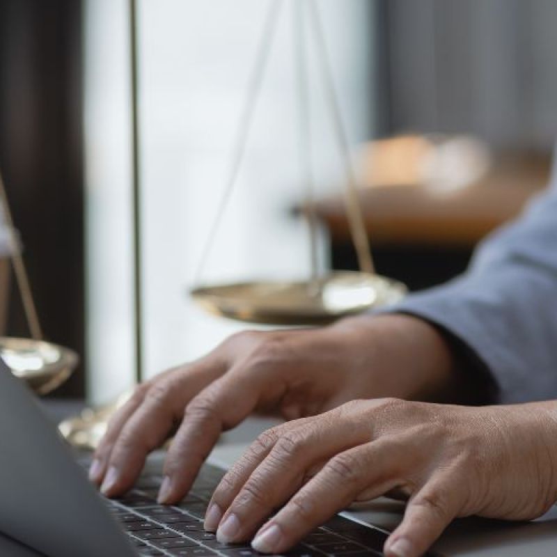 A person types on a laptop while balancing golden scales nearby. The setting is a modern office with soft natural light, suggesting a professional atmosphere.