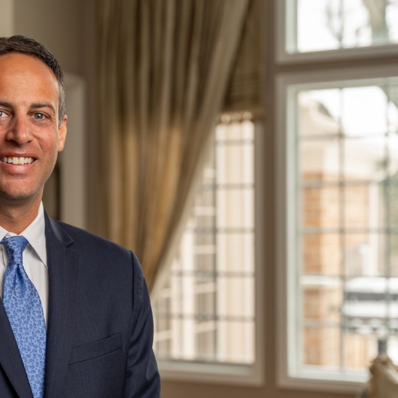 Smiling man in a suit and blue tie, standing inside a well-lit room with large windows in the background.