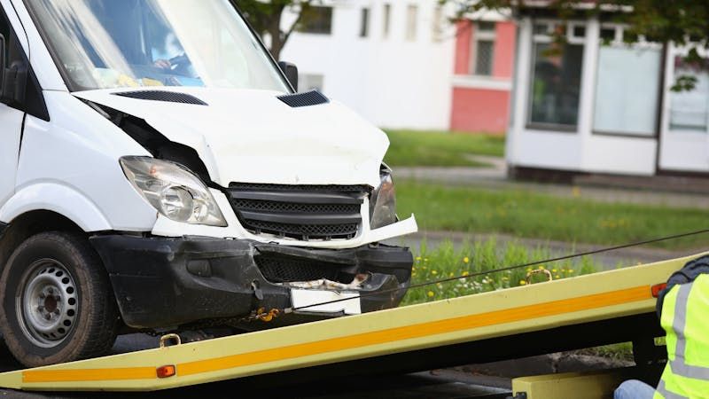 Damaged white van being towed onto a flatbed truck, with a worker in hi-vis gear nearby, in a suburban setting with greenery and buildings in the background.