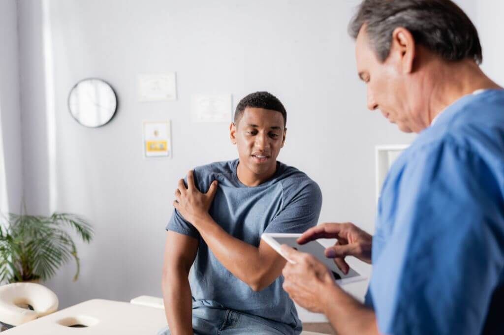 A healthcare professional is using a tablet while examining a young patient's shoulder in a clinical setting with a clock and certificates on the wall.