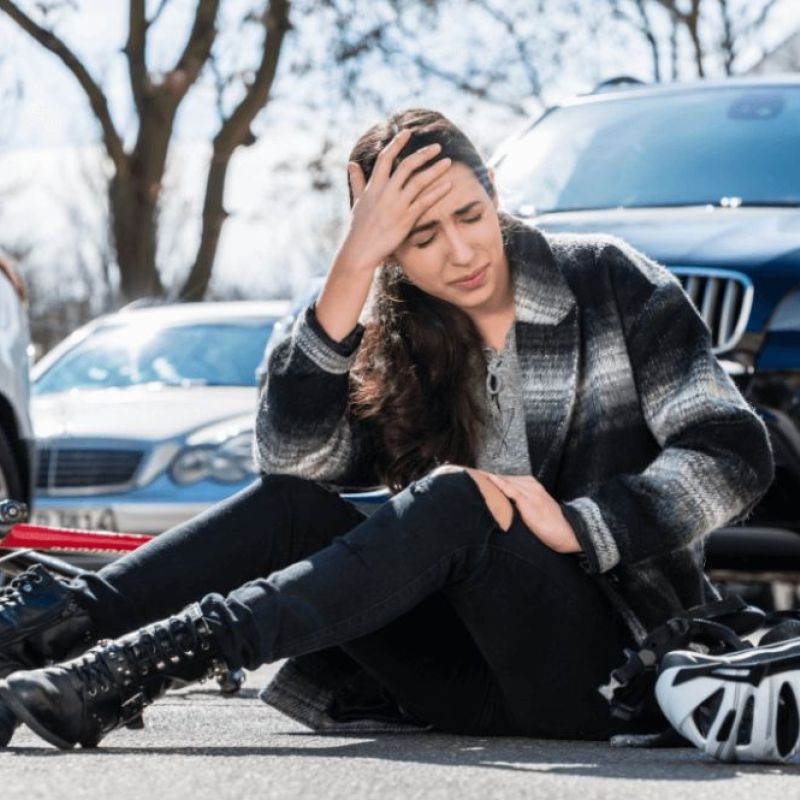 Person sitting on road with bike helmet, looking pained after an accident, surrounded by cars.