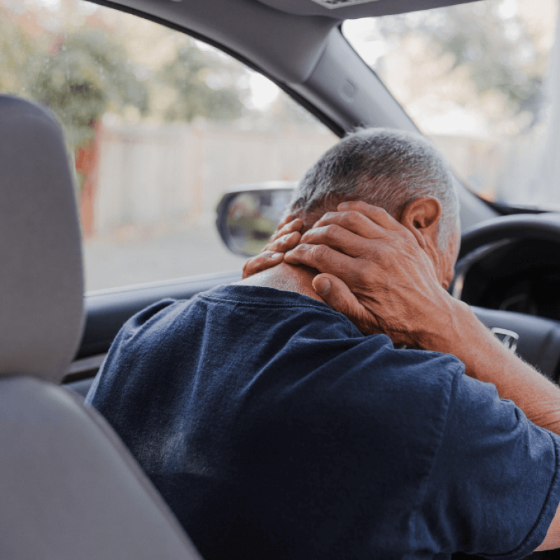 An elderly man sits in a car, gripping his neck with one hand while his other hand rests on the steering wheel, showing signs of discomfort in a blurred outdoor setting.