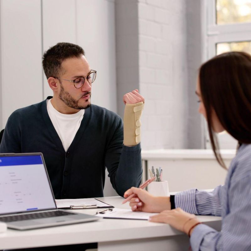 Man with arm cast discusses health insurance claim with a woman in an office setting.