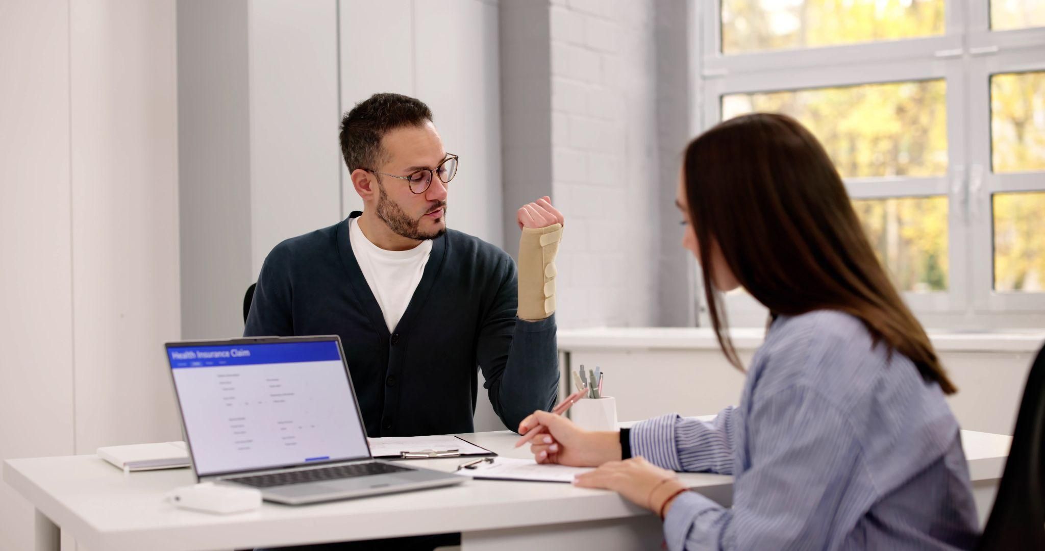 Man with arm cast discusses health insurance claim with a woman in an office setting.