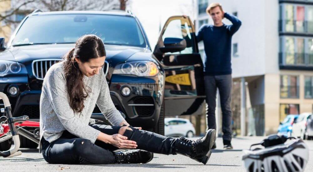 A person sits on the ground by an overturned bicycle in front of a car, suggesting a traffic accident.