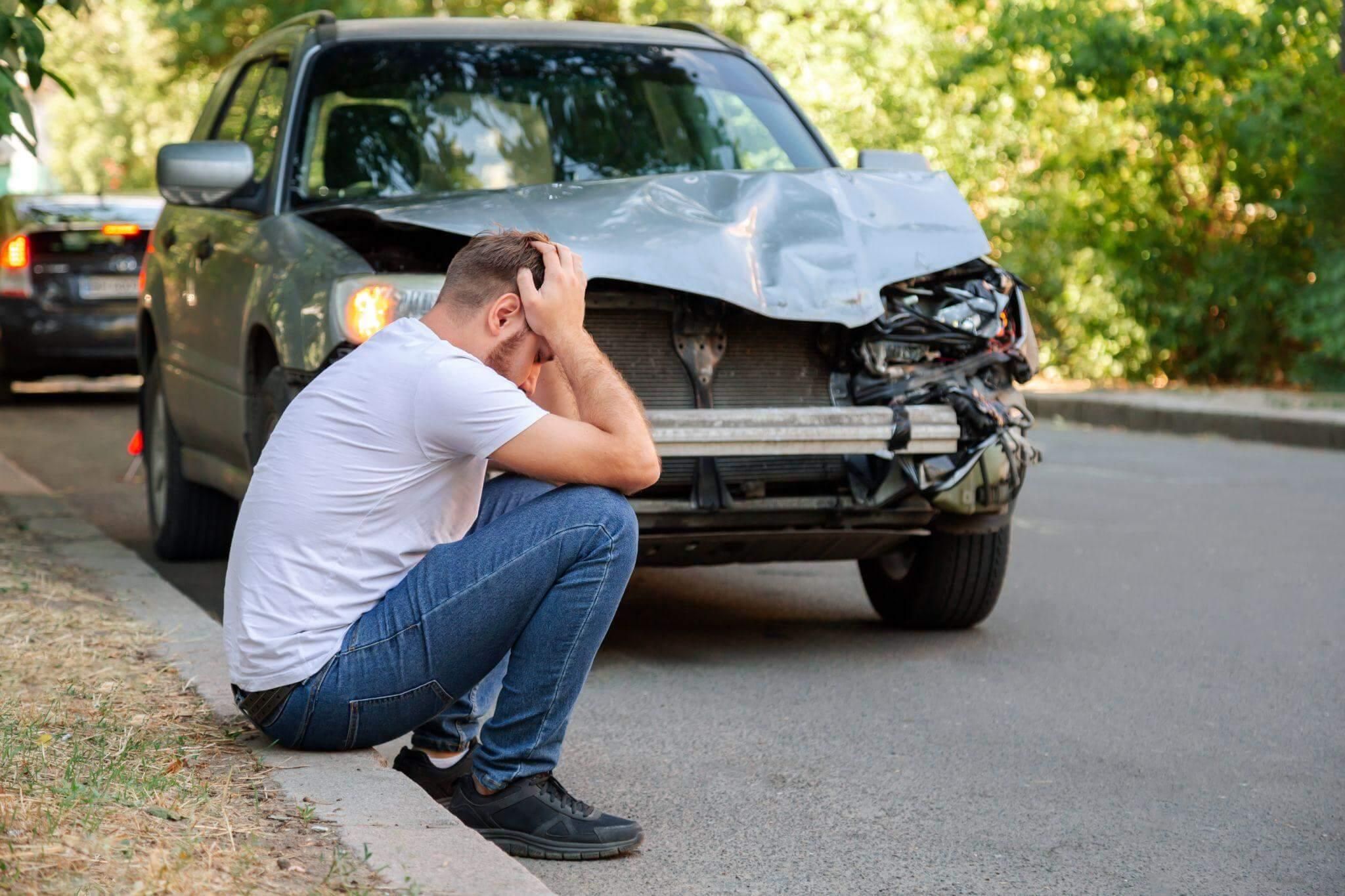 a man sitting on the side of a road next to a car, it seems they were recently involved in a car accident