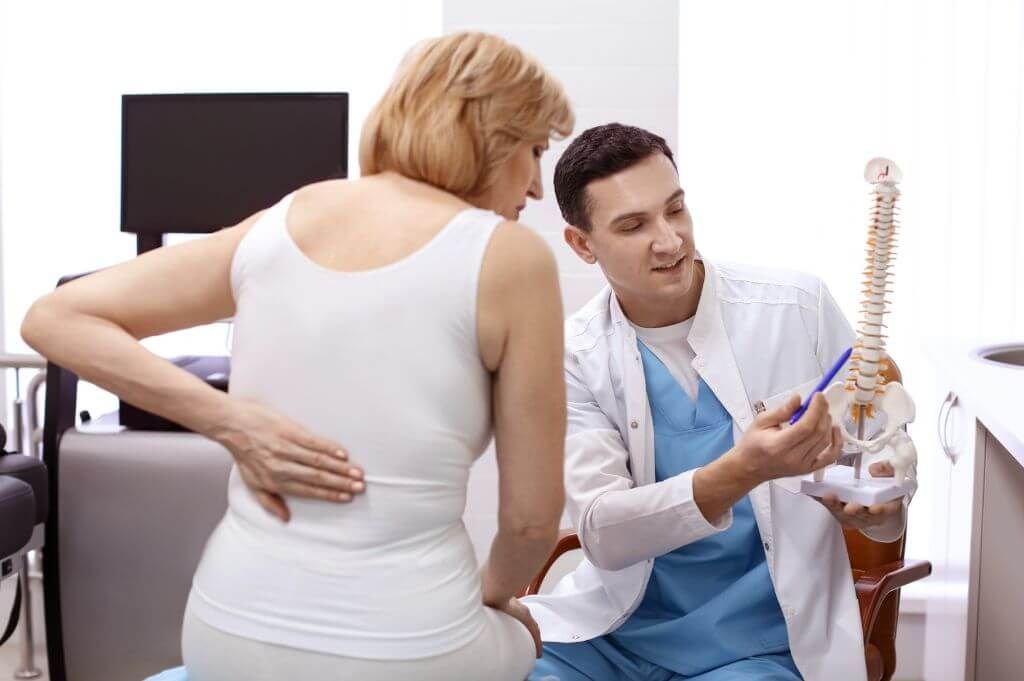 A healthcare professional is explaining a spinal model to a patient touching her back in a clinical setting.