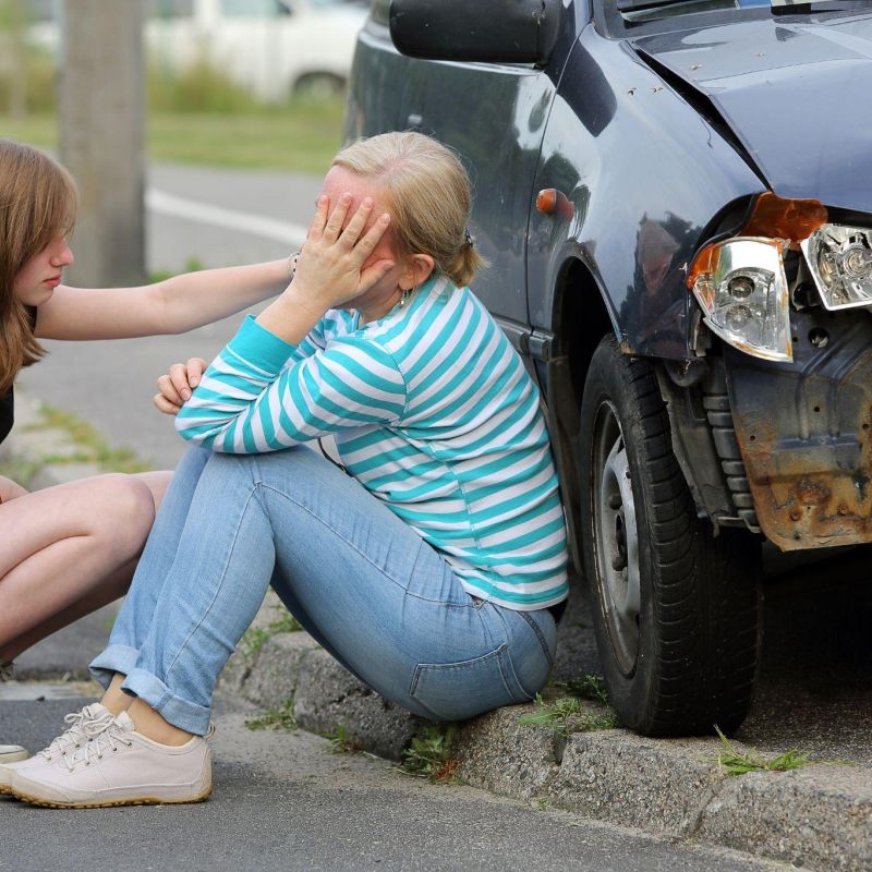 Teen comforting woman after minor car accident, damaged vehicle visible on roadside.