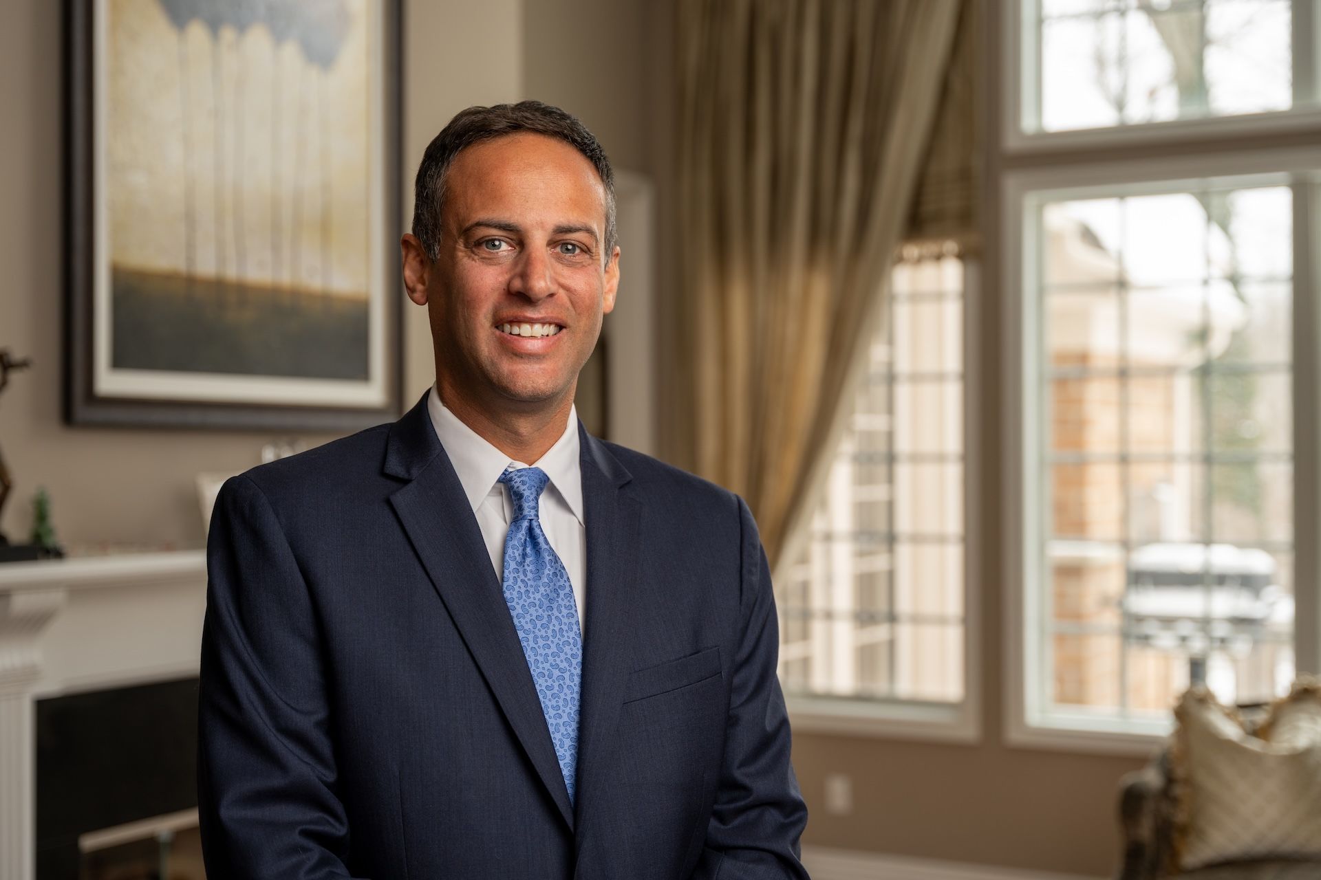Smiling man in a suit and blue tie standing indoors with large windows and artwork in the background.