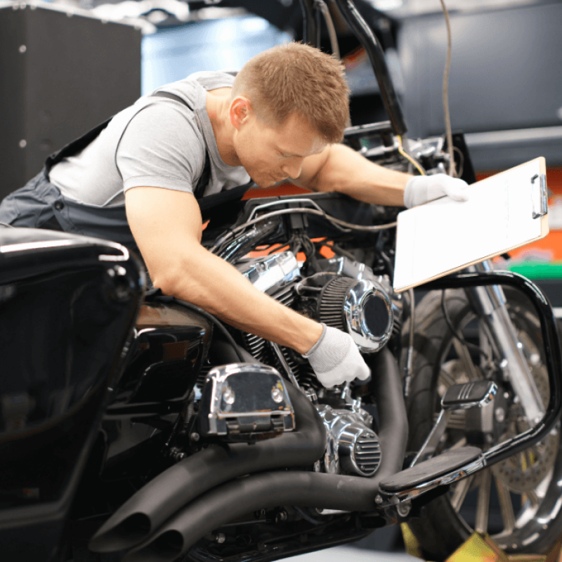 A mechanic works on the engine of a black motorcycle, focused on adjusting components while holding a clipboard. The workspace features tools and equipment in a well-lit, organized garage.