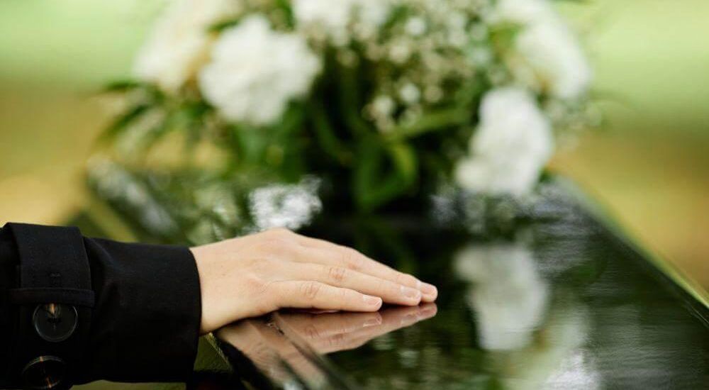 Hand resting on a polished coffin at a funeral, with white-flowered arrangements in the blurred background.