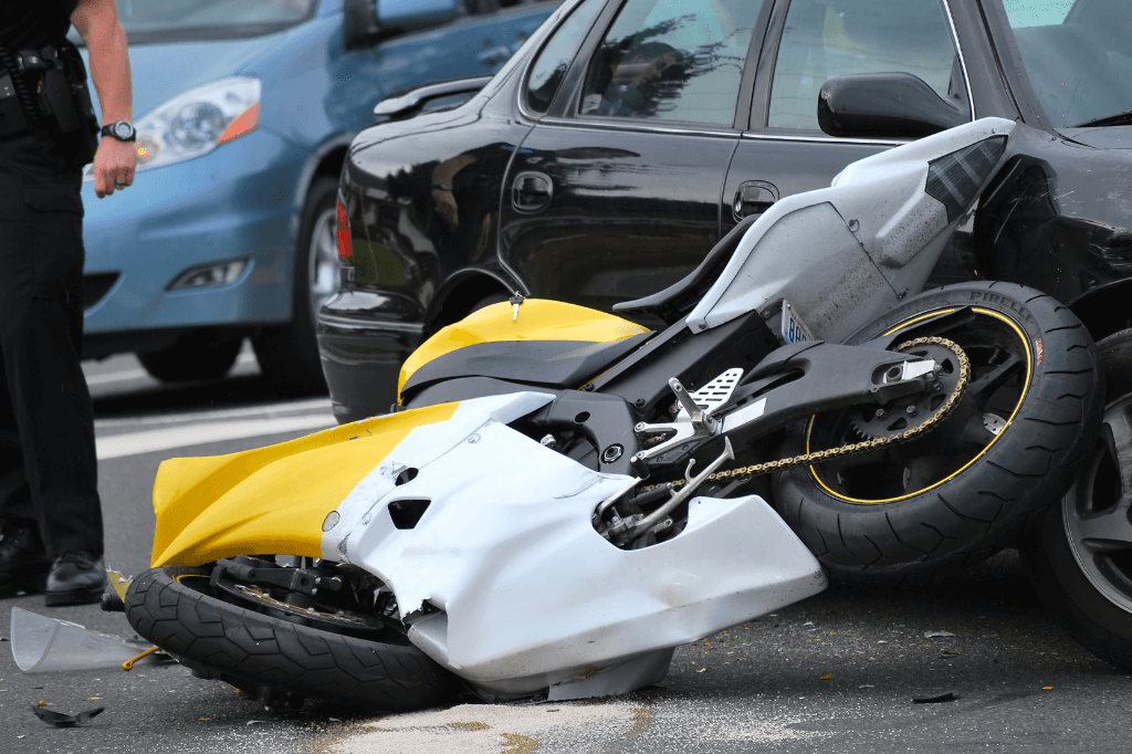 A yellow and white motorcycle lies on its side, partially beneath a black car, suggesting a collision. A police officer stands nearby, assessing the scene on a road.