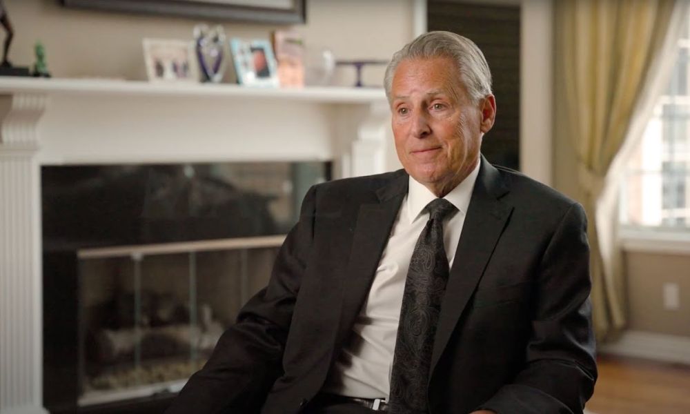 Man in a suit sitting in a well-decorated room, with a fireplace in the background.