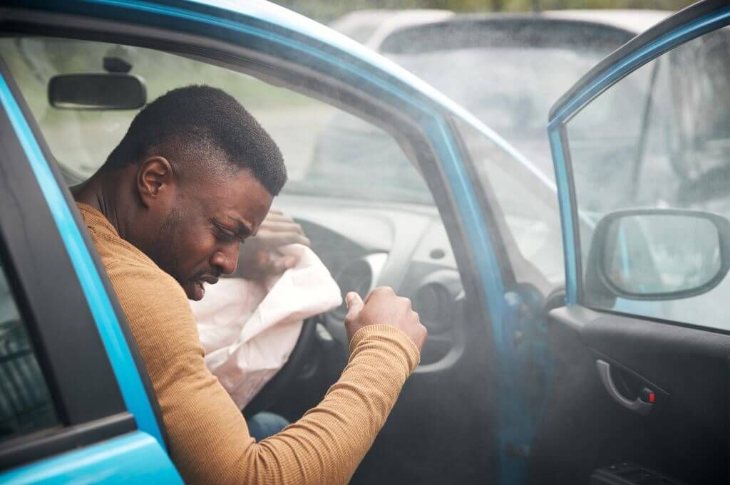 A man sits inside a blue car with an airbag deployed, appearing distressed. Smoke fills the vehicle, suggesting a recent accident in a parking area with blurred cars in the background.