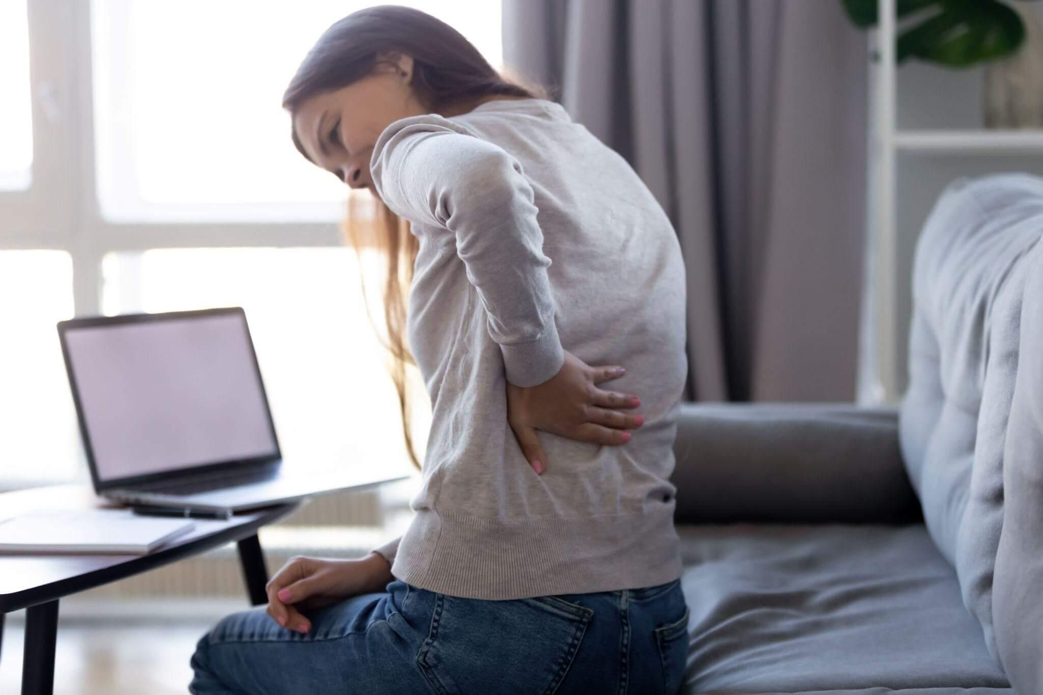 A person holding their lower back in discomfort, seated on a sofa next to a table with a laptop, in a bright and cozy living room.