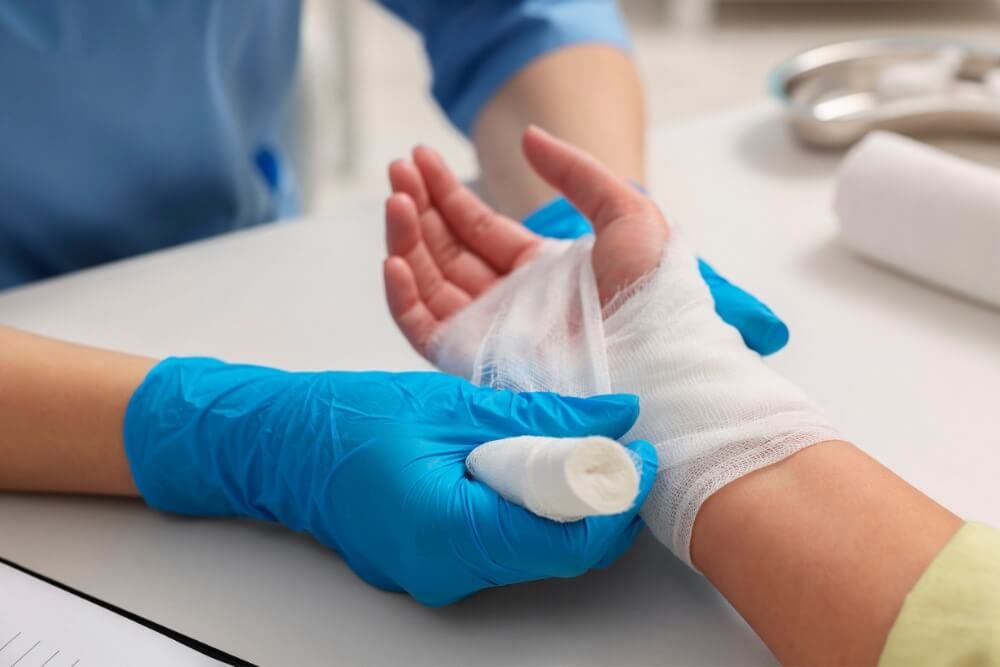 A healthcare worker in blue gloves is wrapping a white bandage around a patient's wrist against a clinical backdrop.