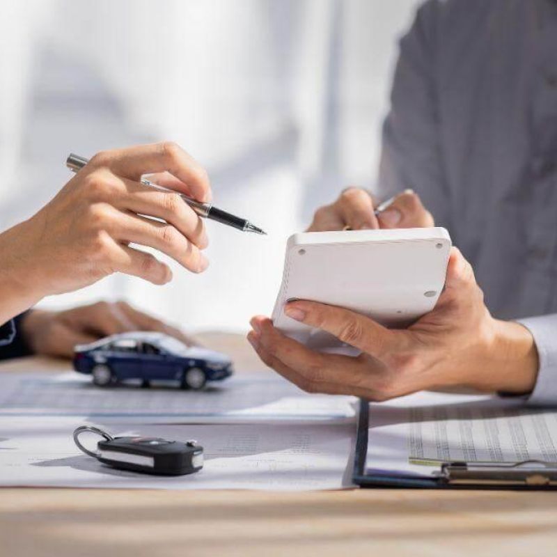 Two professionals reviewing car financing paperwork on a desk, with a small car model, a key fob, and a pen visible.