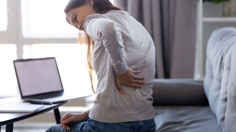 A person holding their lower back in discomfort, seated on a sofa next to a table with a laptop, in a bright and cozy living room.