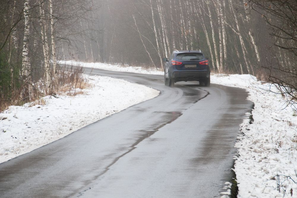 A car driving away on a winding wet road bordered by snow-covered ground and leafless trees.
