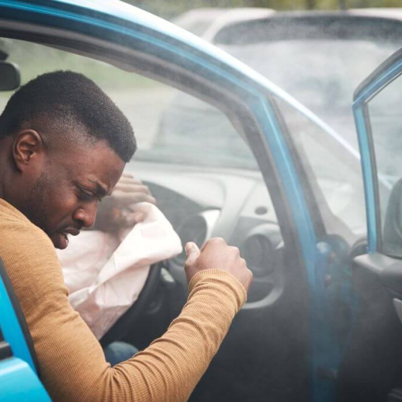 A man sits inside a blue car with an airbag deployed, appearing distressed. Smoke fills the vehicle, suggesting a recent accident in a parking area with blurred cars in the background.