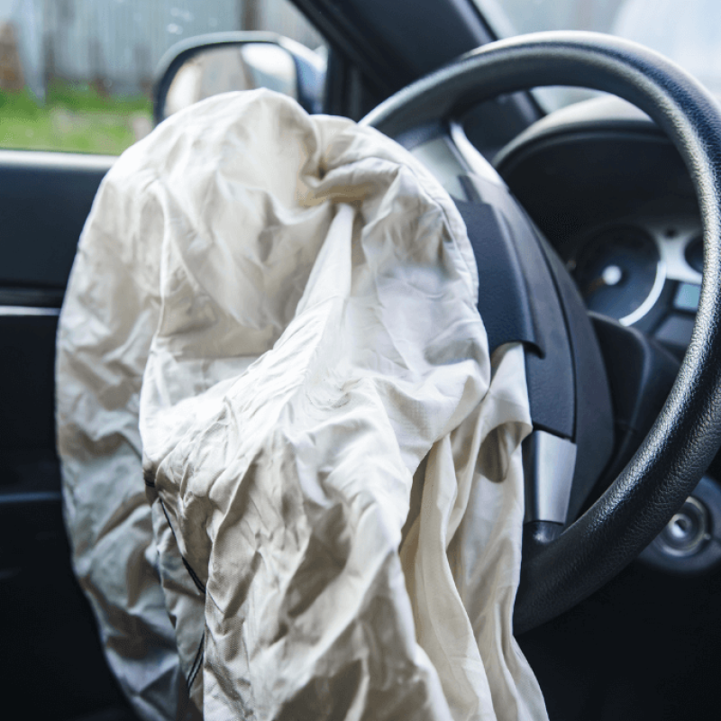 A deployed airbag, crumpled and white, hangs over the steering wheel inside a car. The interior is dimly lit, suggesting an outside environment with blurred greenery visible through the window.