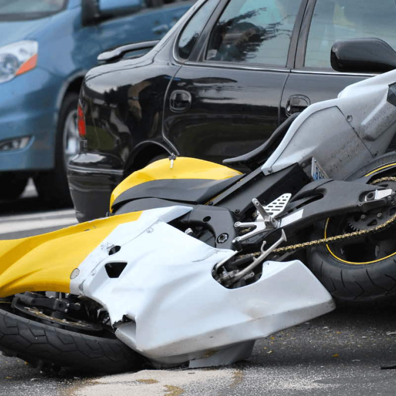 A yellow and white motorcycle lies on its side, partially beneath a black car, suggesting a collision. A police officer stands nearby, assessing the scene on a road.