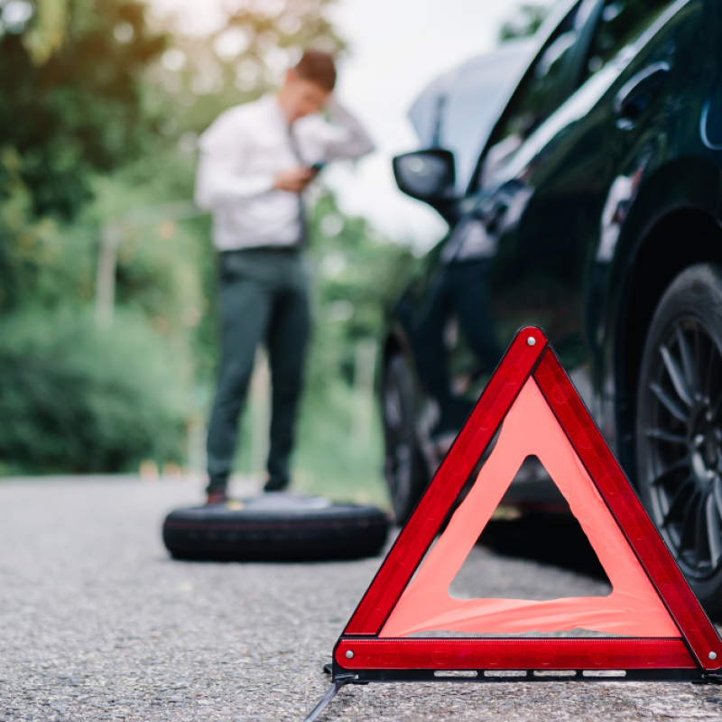 Flat tire roadside with caution triangle and blurred man using phone behind a car.