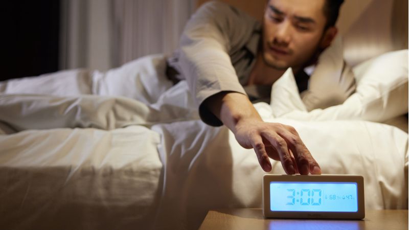 Man reaching for alarm clock at 3:00 AM in bed, illustrating waking up early.