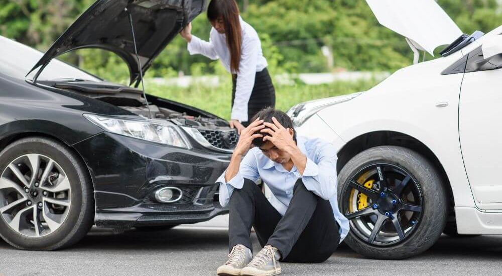 A man sits distraught on the pavement between two cars involved in a minor collision, with a woman standing by one car's open hood. The scene suggests a roadside accident.