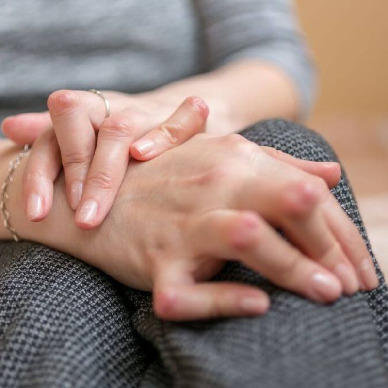 Close-up of a person’s clasped hands resting on their lap, suggesting comfort or contemplation, set against a soft-focus indoor backdrop.