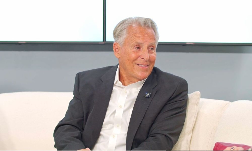 Smiling man in a suit sitting on a couch during an interview, looking to the side.