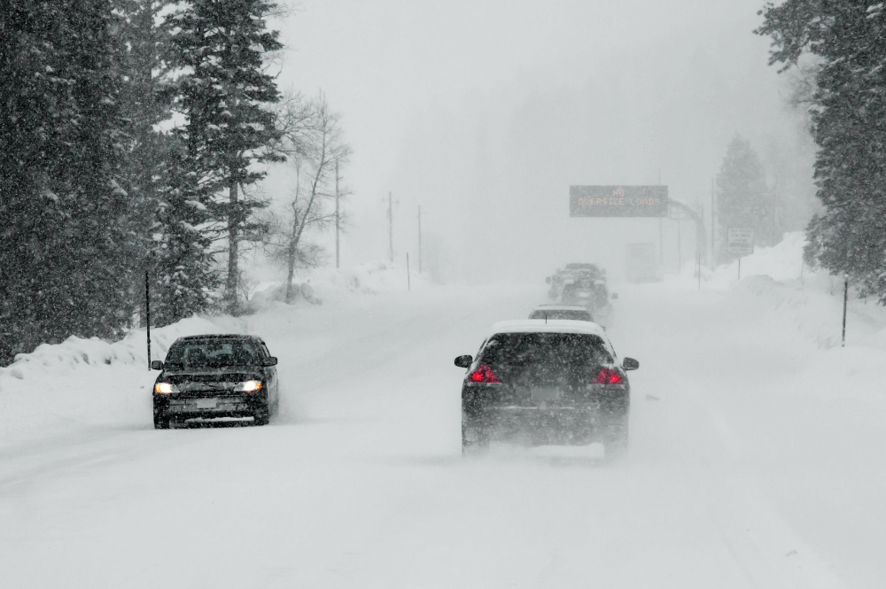 two cars driving in a blizzard on snow covered roads between trees