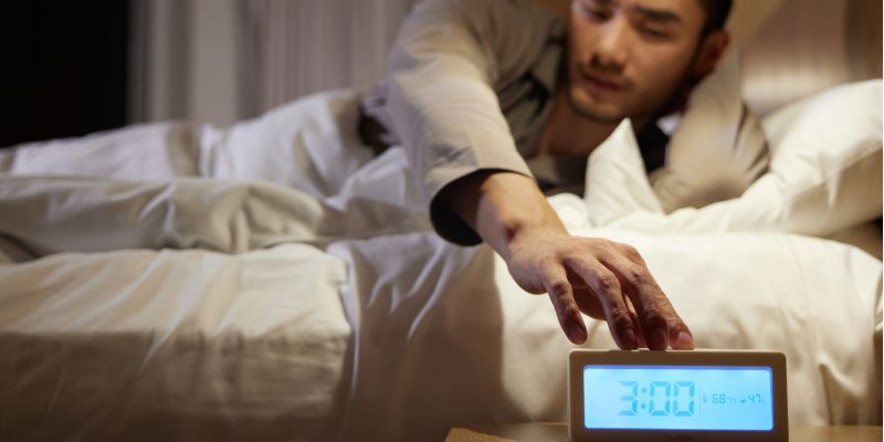 Man reaching for alarm clock at 3:00 AM in bed, illustrating waking up early.