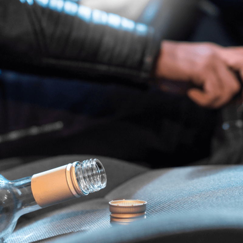 A partially empty glass bottle lies on a car seat, with its cap beside it. A hand grips a gear shift, suggesting the vehicle is in operation.