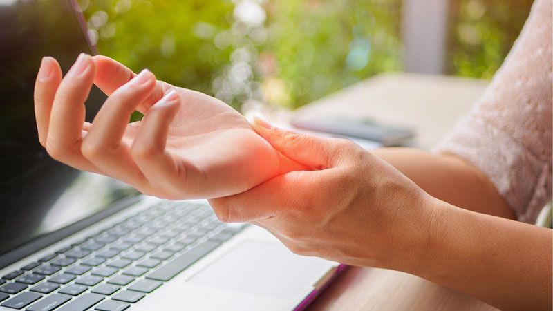 Person holding wrist in pain, sitting at a laptop, indicating potential carpal tunnel or repetitive strain injury.